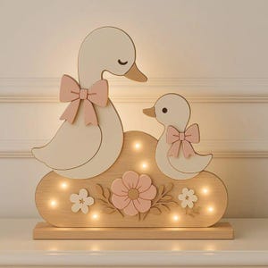 Op de afbeelding: Een lichtgekleurde houten lamp met een grote eend en een klein eendje, beide voorzien van een zachtroze strik. De wolk-vormige voet bevat warm LED-licht en is versierd met uitgesneden houten bloemen en bladeren.