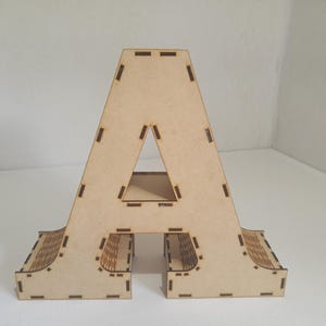 May include: A freestanding wooden letter "A" in a light brown color. The letter has a triangular cutout in the center and decorative cutouts along the bottom edges. The wooden letter is a decorative item.