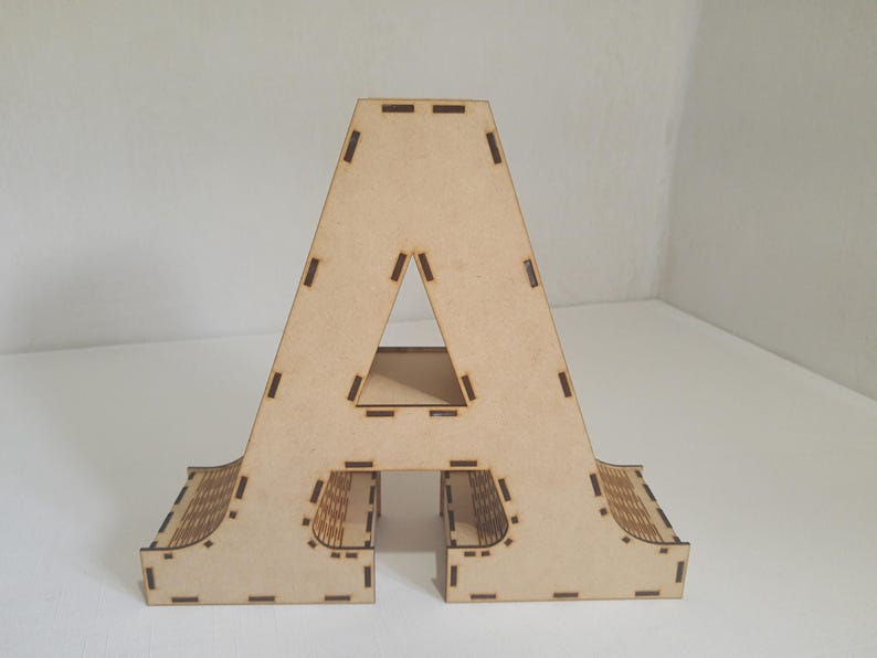 May include: A freestanding wooden letter "A" in a light brown color. The letter has a triangular cutout in the center and decorative cutouts along the bottom edges. The wooden letter is a decorative item.