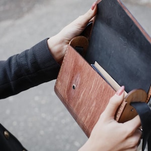 May include: A brown leather satchel with a black interior and a black strap. The satchel is open, revealing a glimpse of the contents inside. The leather has a wood-grain appearance. The satchel is being held open by a person.