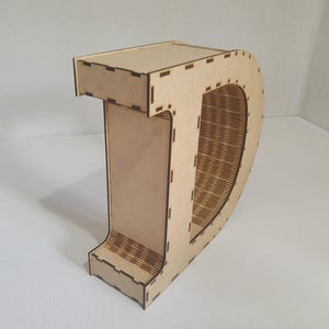 May include: A wooden letter "D" with a decorative design. The letter is made of light brown wood and has a hollowed-out center with a woven pattern. The edges are precisely cut with small rectangular notches.