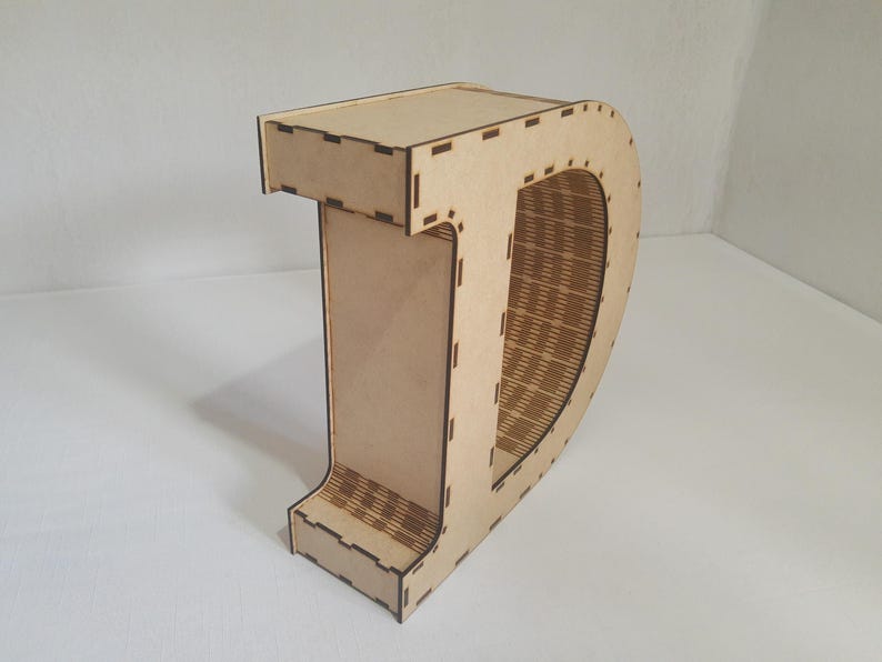 May include: A wooden letter "D" with a decorative design. The letter is made of light brown wood and has a hollowed-out center with a woven pattern. The edges are precisely cut with small rectangular notches.