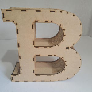 May include: A large, freestanding wooden letter "B" in a light tan color. The letter is constructed from multiple layers of wood, with visible cut lines and slots. The design is simple and modern, suitable for home decor or craft projects.