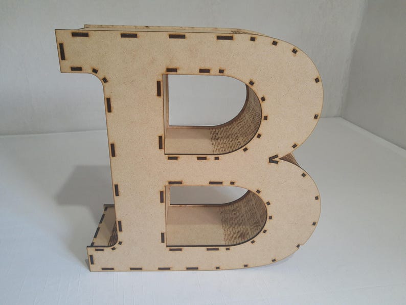 May include: A large, freestanding wooden letter "B" in a light tan color. The letter is constructed from multiple layers of wood, with visible cut lines and slots. The design is simple and modern, suitable for home decor or craft projects.