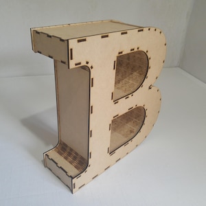 Pode incluir: Uma grande letra "B" de madeira independente com um acabamento de madeira natural. A letra é construída a partir de várias peças de madeira, com linhas de junção visíveis e detalhes recortados. O design inclui duas aberturas arredondadas.