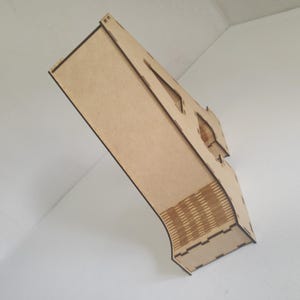 May include: A laser-cut wooden box with a unique architectural design. The box features geometric cutouts and a layered construction, showcasing the natural wood grain. The box is angled against a white background, highlighting its three-dimensional form.