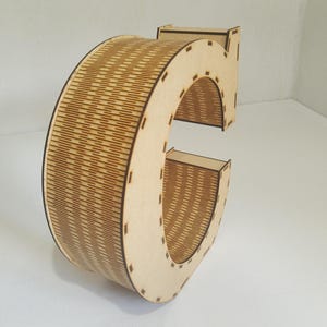 May include: A wooden letter "C" with a natural finish. The letter is constructed from multiple layers of wood, with a detailed, textured pattern on the curved section. The design is suitable for home decor or craft projects.