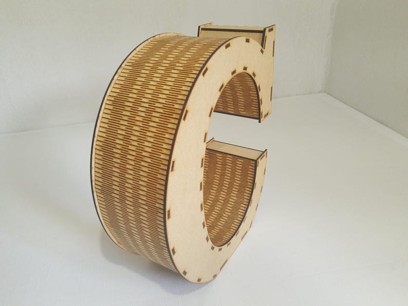 May include: A wooden letter "C" with a natural finish. The letter is constructed from multiple layers of wood, with a detailed, textured pattern on the curved section. The design is suitable for home decor or craft projects.