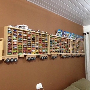 May include: A wooden toy truck-shaped shelf unit mounted on a brown wall, filled with a collection of colourful toy cars. The shelf has multiple compartments to display the cars. Some cars are also displayed on top of the shelf.
