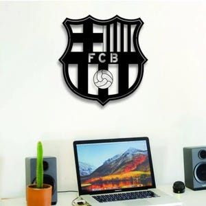 May include: Black metal wall art of the FC Barcelona crest, with the letters "FCB" and a football. It's mounted on a white wall above a desk with a laptop, speakers, and a potted cactus, typical of a home office.