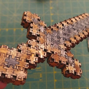 May include: A wooden cross puzzle with interlocking pieces. The cross is made of light and dark brown wood, with some gray pieces. The puzzle is held in a person's hand against a green background.