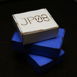 Könnte beinhalten: Drei kleine Pappschachteln übereinander gestapelt. Die unteren beiden sind blau, die obere weiß mit dem Aufdruck "JP08" in Braun. Geeignet für kleine Gegenstände, Schmuck oder Geschenke.