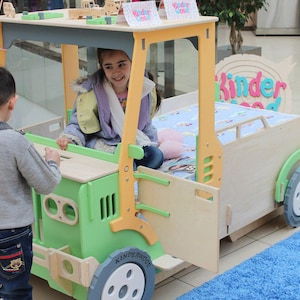 May include: A wooden children's playhouse designed as a car, painted in green, yellow, and natural wood tones. The playhouse features a bed area, wheels, and the text "Kinder Good". Two children are interacting with the playhouse.
