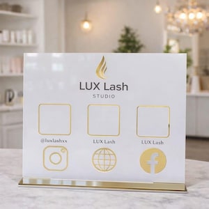 Pode incluir: Um suporte de exibição de acrílico branco com detalhes dourados. O suporte apresenta o texto "LUX Lash STUDIO" e ícones de mídia social para Instagram, um globo e Facebook. O identificador do Instagram é @luxlashxx.