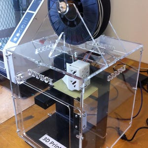 Könnte beinhalten: Ein 3D-Drucker in einem transparenten Acrylgehäuse mit einer schwarzen Filamentrolle auf der Oberseite. Der Drucker verfügt über ein weißes gedrucktes Objekt auf der Druckplatte und ein weißes Etikett mit der Aufschrift 3D Printer 1.