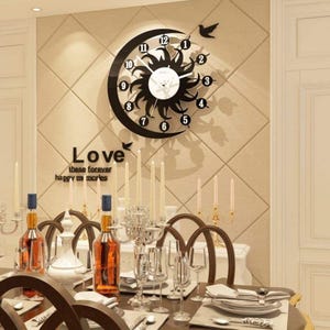 May include: A black and white wall clock with a sun and moon design, featuring the numbers 1 through 12. The clock is mounted on a beige wall with the word "Love" and the phrase "these forever happy memories" below it. The image also shows a dining table with bottles and glasses.
