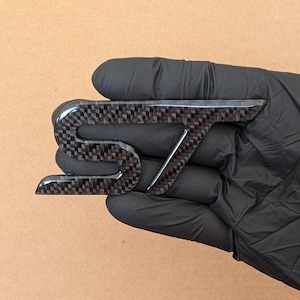 Op de afbeelding: Een zwart en grijs koolstofvezel embleem met de letters ST in een gestileerd lettertype. Het embleem heeft een glanzende afwerking met een zichtbaar geweven patroon over het oppervlak van de letters, wat zorgt voor een textuur op dit autobadge.