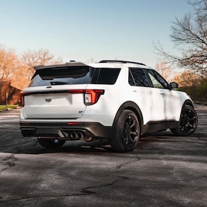 May include: A white Ford Explorer ST SUV viewed from the rear three-quarter angle. The vehicle features black alloy wheels, a rear spoiler, quad exhaust tips, and a black ST badge on the tailgate. The taillights are red and clear with a horizontal design.