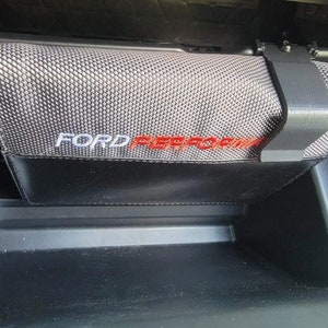 Pode incluir: Uma caixa de armazenamento preta coberta de tecido com uma parte superior de malha cinzenta e um clipe de plástico preto. A caixa tem as palavras "FORD PERFORMANCE" impressas em branco e vermelho.