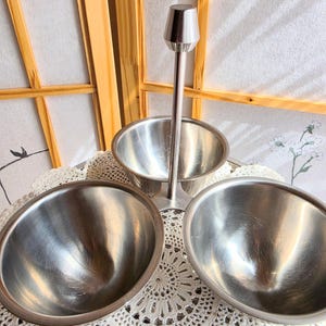 May include: A stainless steel serving set with three bowls. The set includes two oval bowls and one bowl on a tall stand with a conical top. The bowls are arranged on a white lace tablecloth.