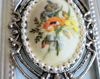 May include: A silver and white cameo brooch with a floral design. The cameo features a bouquet of flowers in shades of orange, yellow, and blue.