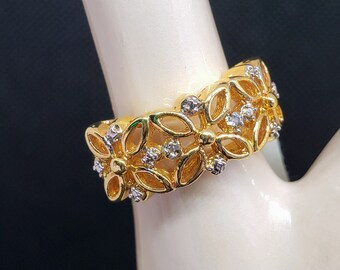 May include: A gold-toned ring featuring a floral design with openwork petals. The ring is adorned with small, clear gemstones. The ring is on a white surface against a dark background.