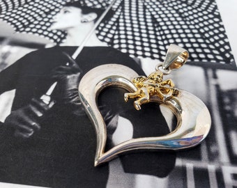 May include: Silver heart-shaped pendant with a gold cherub holding an arrow. The pendant is on a black and white background.