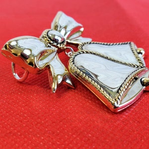May include: A silver and gold-toned brooch featuring a white bell with a gold trim and a bow with a gold center.