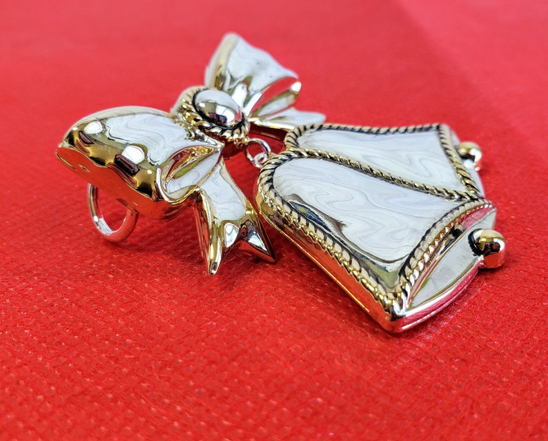 May include: A silver and gold-toned brooch featuring a white bell with a gold trim and a bow with a gold center.