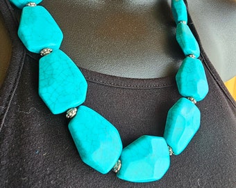 May include: A turquoise statement necklace featuring large, irregularly shaped beads. The beads are a vibrant blue-green color, connected by small silver-toned spacers. The necklace is displayed on a black mannequin.