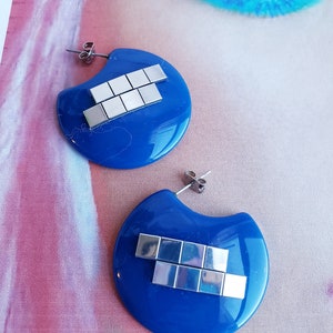 May include: A pair of blue resin earrings with silver geometric shapes. The earrings are shaped like a crescent moon.