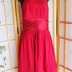 Op de afbeelding: Een felrode halterjurk met een geplooid lijfje en een getailleerde taille. De jurk heeft een zwierige rok en wordt gepresenteerd op een zwarte mannequin. Deze elegante jurk is geschikt voor speciale gelegenheden.