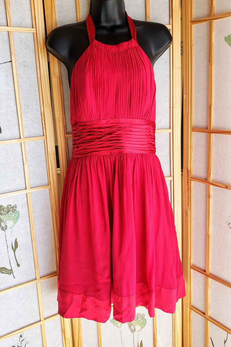 Op de afbeelding: Een felrode halterjurk met een geplooid lijfje en een getailleerde taille. De jurk heeft een zwierige rok en wordt gepresenteerd op een zwarte mannequin. Deze elegante jurk is geschikt voor speciale gelegenheden.