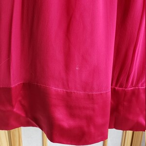 Op de afbeelding: Close-up van een levendige fuchsia-kleurige stof, mogelijk een jurk of rok. Het materiaal heeft een gladde textuur met een contrasterende satijnen zoom. De afbeelding benadrukt de kleur en de val van de stof.