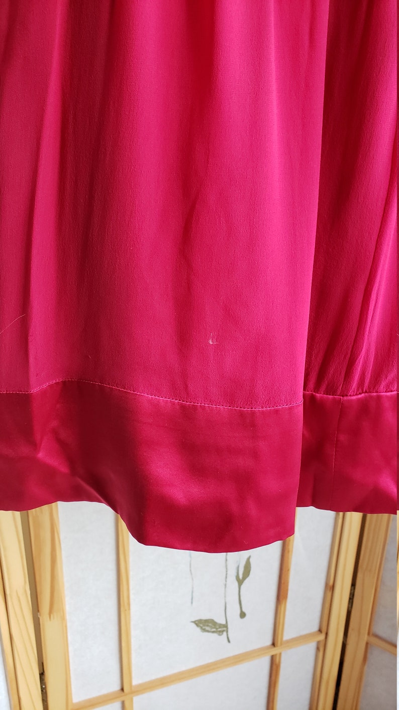 Op de afbeelding: Close-up van een levendige fuchsia-kleurige stof, mogelijk een jurk of rok. Het materiaal heeft een gladde textuur met een contrasterende satijnen zoom. De afbeelding benadrukt de kleur en de val van de stof.