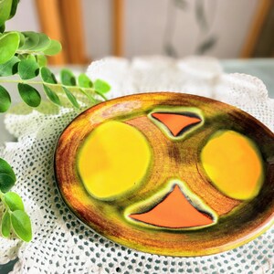 May include: A ceramic plate with a yellow, orange, and green abstract design. The plate has a brown rim and is decorated with a swirling pattern.