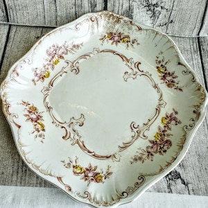 May include: A white ceramic serving platter with a scalloped edge and a floral design in shades of brown and yellow. The design features intricate scrollwork and delicate floral motifs.