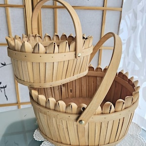 May include: Two light brown, handcrafted wooden baskets with arched handles. The baskets have a decorative scalloped rim and are constructed with vertical wooden slats. The baskets are stacked on top of each other.