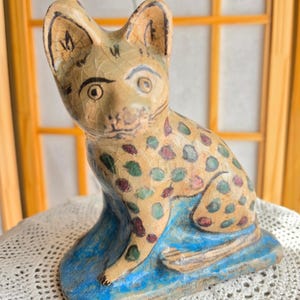 May include: A ceramic cat figurine with a tan body and blue base. The cat has black markings on its ears and face, and colorful polka dots on its body. The figurine is sitting on a white lace surface.