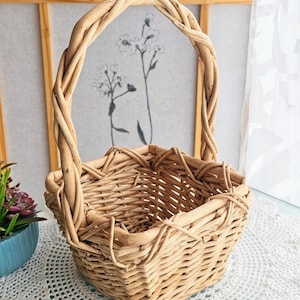 May include: A natural woven basket with a sturdy handle. The basket is square-shaped with a woven design and a tall, arched handle. The basket is made of light brown wicker and sits on a white lace tablecloth.