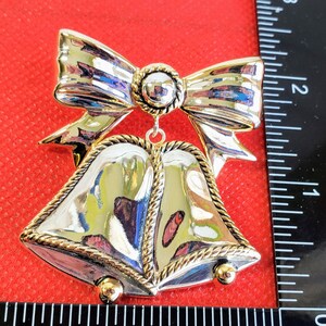 May include: Silver and gold Christmas ornament featuring two bells with a bow on top. The bells have a rope-like design around the edges.