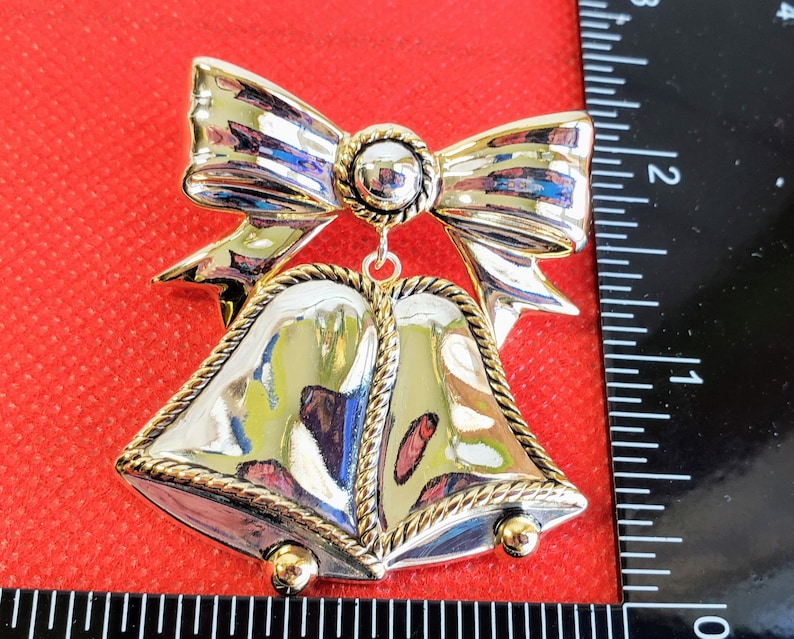 May include: Silver and gold Christmas ornament featuring two bells with a bow on top. The bells have a rope-like design around the edges.
