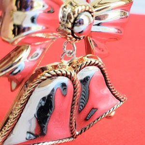 May include: A silver and gold Christmas ornament featuring two bells with a bow on top. The bells are decorated with a gold rope design.