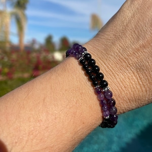 Könnte beinhalten: Ein mehrreihiges Armband mit zwei Strängen aus Perlen. Ein Strang besteht aus dunkelvioletten Amethystperlen, der andere aus schwarzen Onyxperlen. Das Armband wird am Handgelenk getragen.