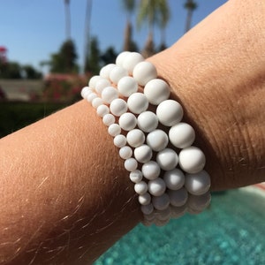 Puede incluir: Un conjunto de cuatro pulseras de cuentas blancas apiladas en la muñeca de una persona. Las pulseras están hechas de cuentas lisas y redondas de diferentes tamaños.