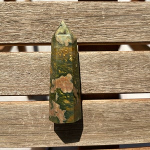 Puede incluir: Una punta de piedra pulida de color verde, marrón y dorado con una parte superior puntiaguda. La piedra tiene un patrón único de remolinos y manchas.