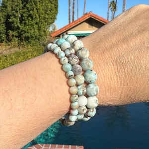 May include: Three stacked bracelets made of light blue and white gemstone beads. The bracelets are worn on a person's wrist.