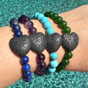 May include: Three bracelets stacked on a wrist. The top bracelet is green, the middle bracelet is turquoise, and the bottom bracelet is blue. Each bracelet has a black lava rock heart charm.