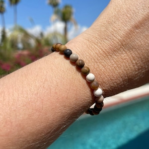 May include: A brown, white, and black beaded bracelet with a natural stone look. The bracelet is worn on a person's wrist.