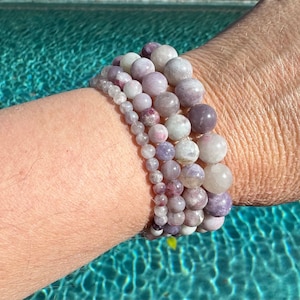 May include: A stack of five bracelets made of light purple and white gemstone beads. The bracelets are worn on a person's wrist.